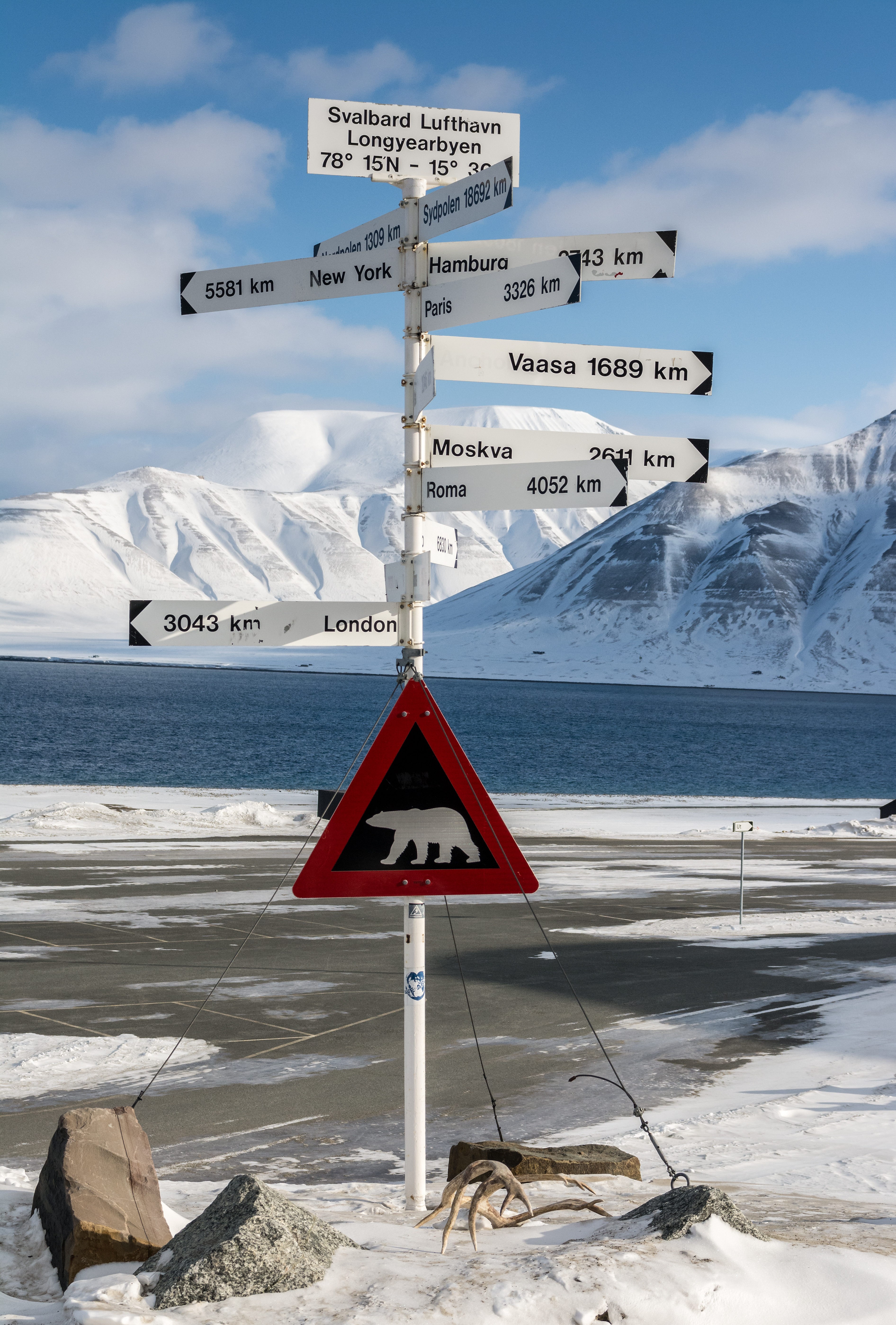 Polar bear crossing with signs of distances to major cities around the world. Image shows white mountains in the background in this arctic climate. the signs are tied to the ground so the wind doesn't blow them away. The signs are taken against a clear blue sky. the signs is located outside Logyearbyen airport. (Getty)